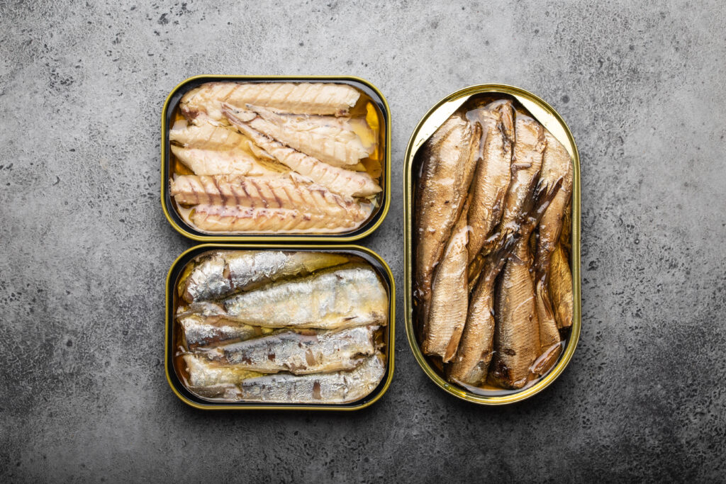 Assorted canned fish in a tin over gray concrete background: sardine, smoked sardine, mackerel.